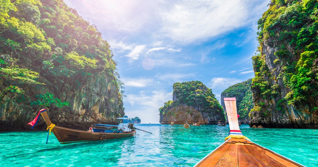 Long tailed boats are anchored lapped by green waters with a limestone rocky outcrop in the distance